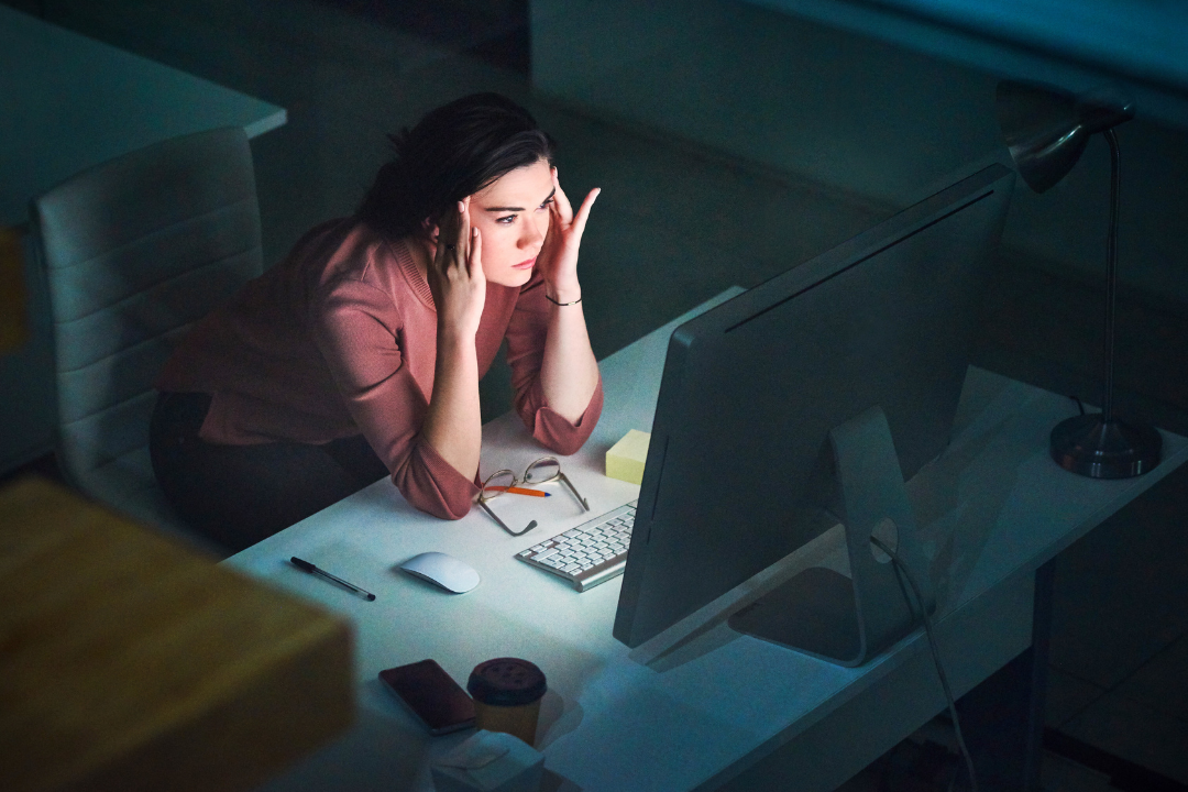 A woman sitting at her desk late at night holding her head while staring at a computer screen, showing common but often missed symptoms of stress.