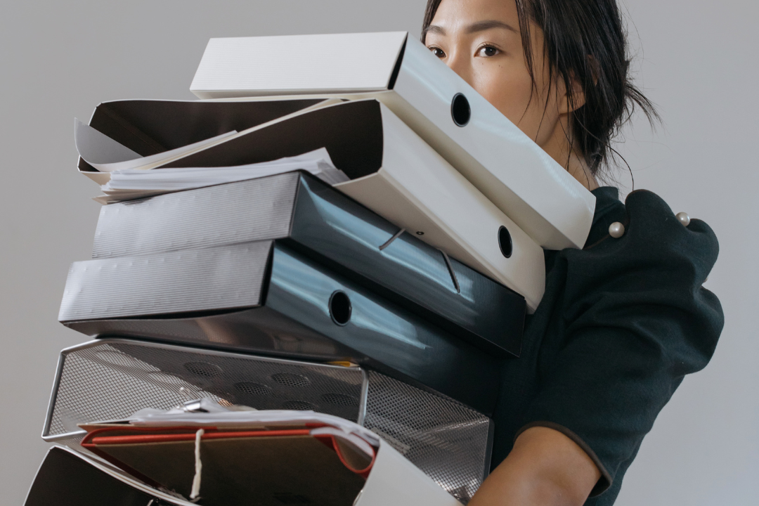 6 Signs You're Working Too Hard And It's Costing Your Success Overwhelmed professional carrying tall stack of files and binders representing signs of working too hard and burnout